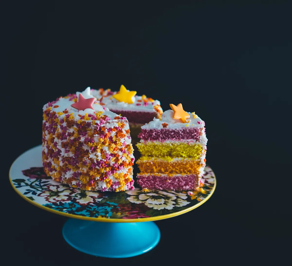Tarta cubierta de virutas de colores, cortada para enseñar seis capas de bizcocho de colores, sobre un pie de tarta azul