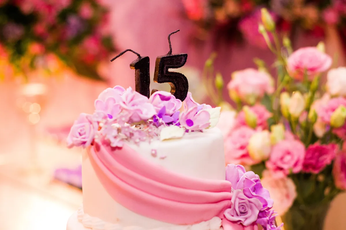 Tarta blanca con una banda de fondant rosa, flores lilas y un 15 en lo alto, rodeada de flores